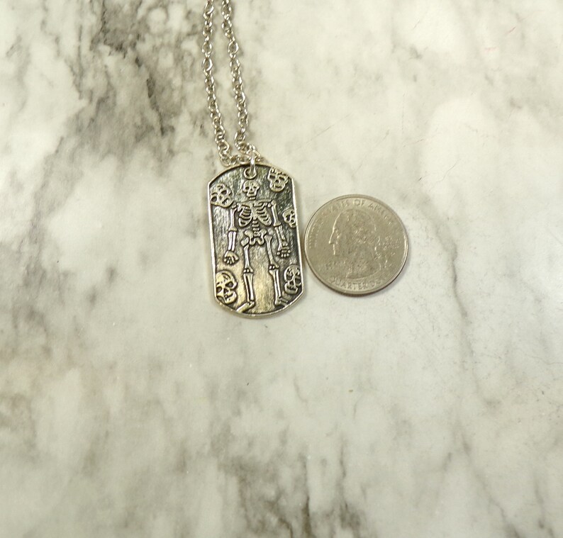 May include: Silver necklace with a skeleton pendant surrounded by skulls. The pendant is shaped like a dog tag.