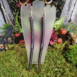 May include: A pair of dangle earrings featuring long, dark brown worm-shaped charms. The earrings are displayed on a white jewelry stand.