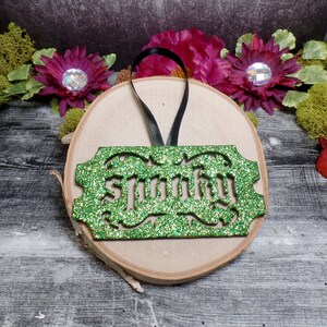 Puede incluir: Adorno verde brillante con la palabra "Spunky" recortada en una fuente decorativa. El adorno rectangular tiene un borde decorativo y una cinta negra para colgar. Descansa sobre una rodaja de madera con detalles florales.