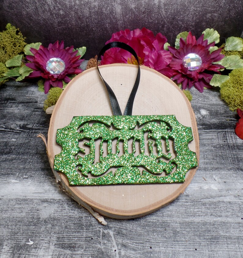 Puede incluir: Adorno verde brillante con la palabra "Spunky" recortada en una fuente decorativa. El adorno rectangular tiene un borde decorativo y una cinta negra para colgar. Descansa sobre una rodaja de madera con detalles florales.