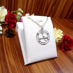 May include: A silver necklace with a circular pendant featuring a sigil design. The pendant is on a white display stand and surrounded by flowers.