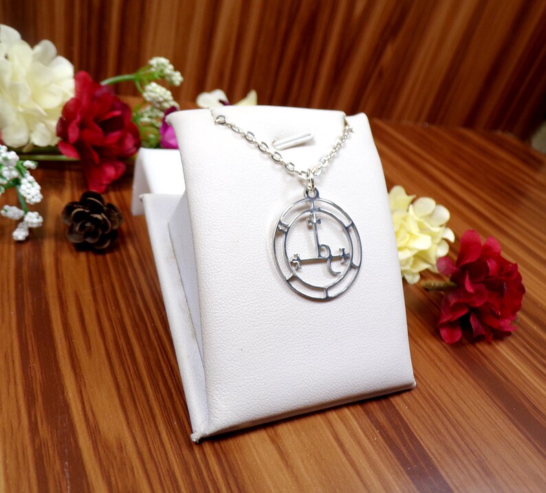 May include: A silver necklace with a circular pendant featuring a sigil design. The pendant is on a white display stand and surrounded by flowers.