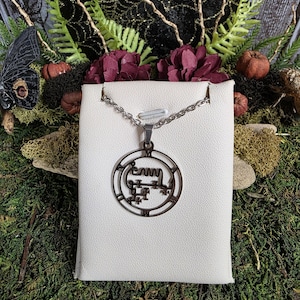 May include: A silver-colored pendant necklace featuring a circular pendant with an intricate, stylized design. The pendant hangs from a delicate chain and is displayed on a white surface, surrounded by decorative elements including flowers and greenery.