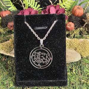May include: A silver-colored pendant necklace with a circular design featuring intricate patterns and symbols. The pendant hangs from a delicate chain and is displayed on a black velvet surface, with decorative elements in the background.