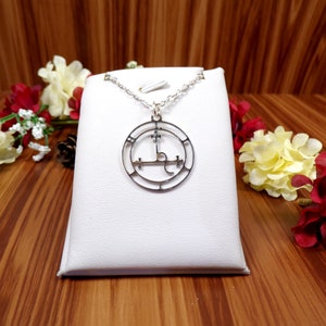 May include: A silver necklace with a circular pendant featuring a sigil design. The pendant is on a white display stand.