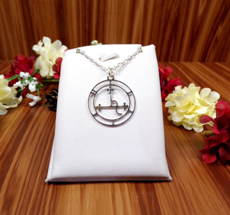 May include: A silver necklace with a circular pendant featuring a sigil design. The pendant is on a white display stand.