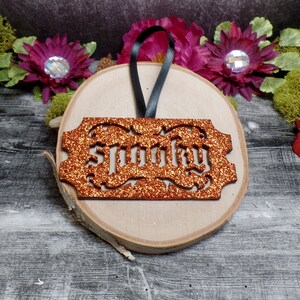 May include: An orange glitter "Spooky" ornament with a black ribbon, presented on a wooden slice. The ornament features a decorative border and is set against a backdrop of flowers and greenery, ideal for Halloween.