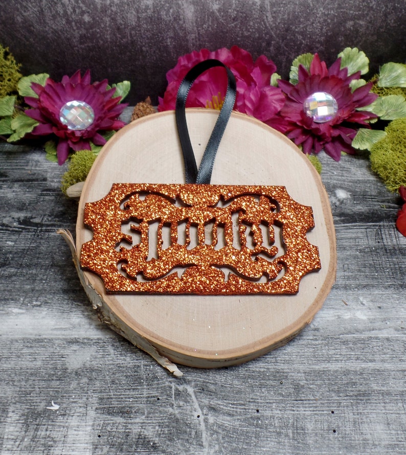 May include: An orange glitter "Spooky" ornament with a black ribbon, presented on a wooden slice. The ornament features a decorative border and is set against a backdrop of flowers and greenery, ideal for Halloween.