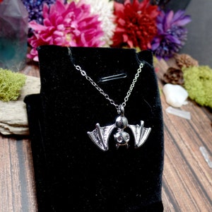 Pu&ograve; includere: Una collana con un ciondolo a forma di pipistrello argentato su uno sfondo di velluto nero. Il pipistrello ha un design dettagliato con le ali spiegate.