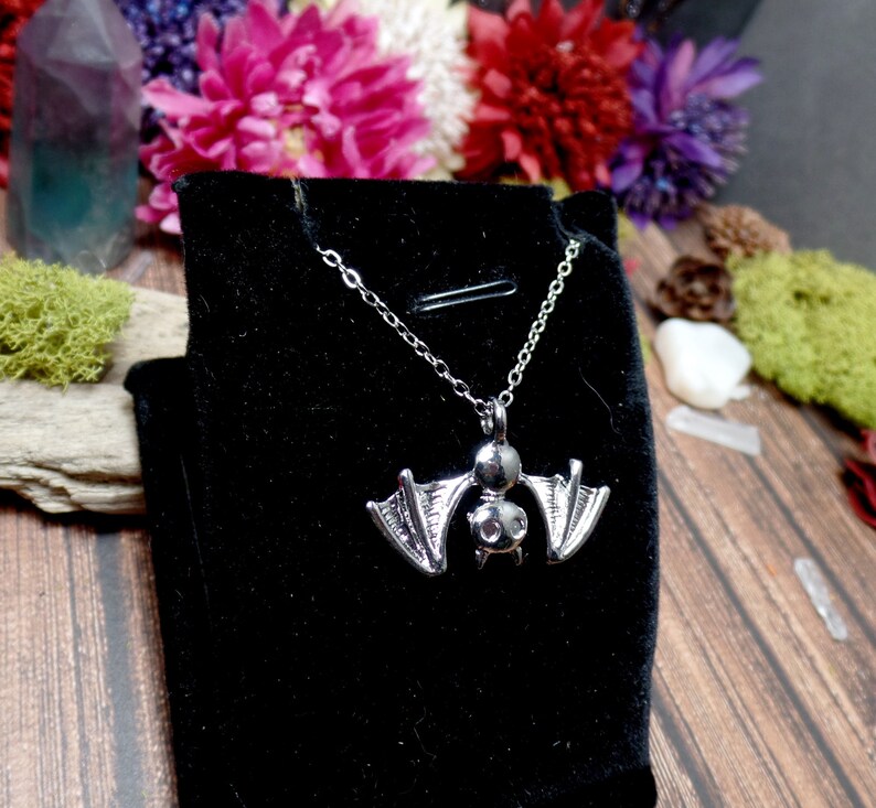 Pu&ograve; includere: Una collana con un ciondolo a forma di pipistrello argentato su uno sfondo di velluto nero. Il pipistrello ha un design dettagliato con le ali spiegate.
