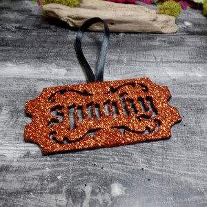 May include: Orange glittery ornament with the word "Spooky" cut out. The ornament has a black ribbon for hanging. The background is a grey wooden surface with decorative elements.
