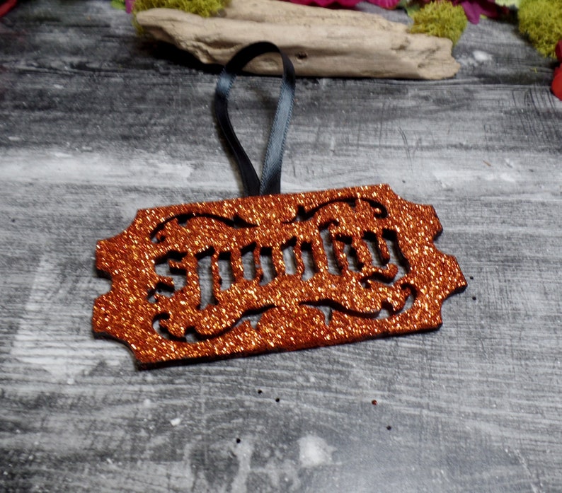 May include: Orange glittery ornament with the word "Spooky" cut out. The ornament has a black ribbon for hanging. The background is a grey wooden surface with decorative elements.