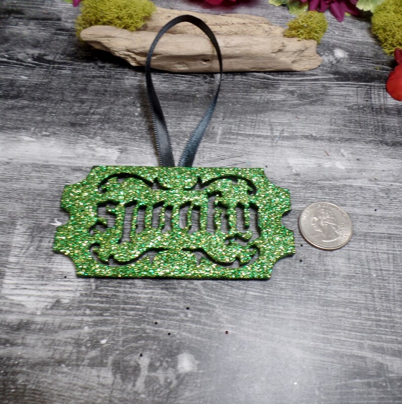 Puede incluir: Un adorno verde brillante con la palabra "Smirky" recortada. El adorno tiene una cinta negra para colgar. Una moneda est&aacute; al lado del adorno para comparar el tama&ntilde;o. El adorno est&aacute; sobre una superficie de madera gris.