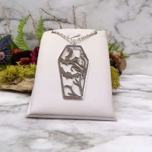 Pu&ograve; includere: Una collana d'argento con un ciondolo a forma di bara che presenta un design di taglio dettagliato di un pipistrello e viti. Il ciondolo &egrave; esposto su un espositore per gioielli bianco.