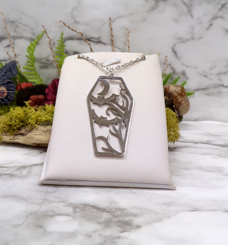 Pu&ograve; includere: Una collana d'argento con un ciondolo a forma di bara che presenta un design di taglio dettagliato di un pipistrello e viti. Il ciondolo &egrave; esposto su un espositore per gioielli bianco.