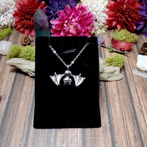 Pu&ograve; includere: Una collana con un ciondolo a forma di pipistrello argentato su uno sfondo di velluto nero. La collana &egrave; esposta su una superficie di legno con fiori e cristalli.