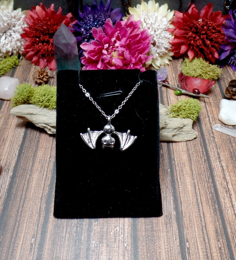 Pu&ograve; includere: Una collana con un ciondolo a forma di pipistrello argentato su uno sfondo di velluto nero. La collana &egrave; esposta su una superficie di legno con fiori e cristalli.