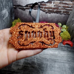 May include: An orange glitter ornament with the word "Smutty" cut out in a decorative font. It has a dark grey ribbon for hanging. The background features brick and wood textures.