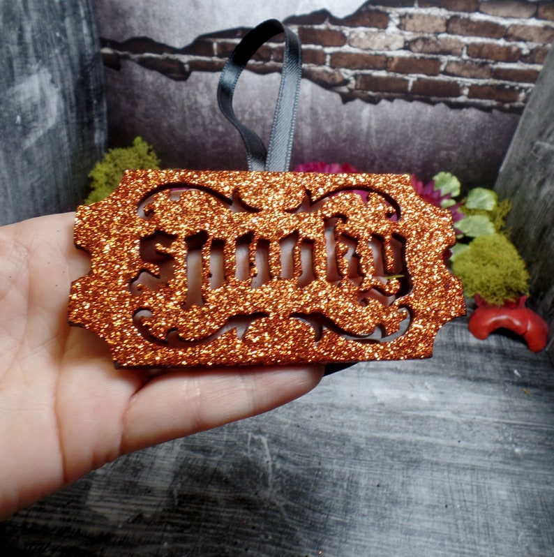 May include: An orange glitter ornament with the word "Smutty" cut out in a decorative font. It has a dark grey ribbon for hanging. The background features brick and wood textures.