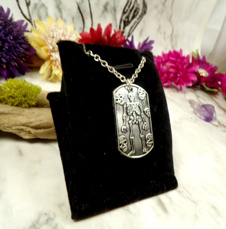May include: A silver skeleton pendant necklace with a skull design. The pendant is on a black velvet background.