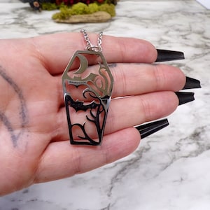 Pu&ograve; includere: Una collana argentata con un grande ciondolo traforato a forma di bara. Il ciondolo presenta un pipistrello, una luna crescente e un motivo a vite.
