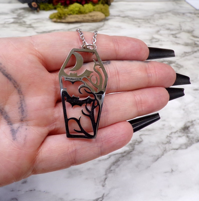 Pu&ograve; includere: Una collana argentata con un grande ciondolo traforato a forma di bara. Il ciondolo presenta un pipistrello, una luna crescente e un motivo a vite.
