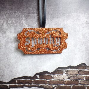 May include: An orange glittery ornament with the word "lucky" cut out in a decorative font. The ornament is rectangular with ornate edges and is suspended by a grey ribbon. The background is a textured grey wall with a brick pattern.