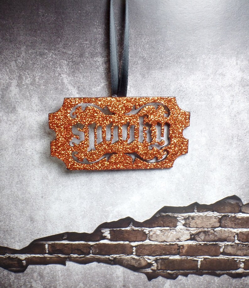 May include: An orange glittery ornament with the word "lucky" cut out in a decorative font. The ornament is rectangular with ornate edges and is suspended by a grey ribbon. The background is a textured grey wall with a brick pattern.