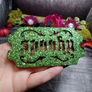 Puede incluir: Un adorno verde brillante con la palabra "Smitty" recortada en una fuente decorativa. El adorno tiene una cinta negra para colgar y est&aacute; rodeado de flores y vegetaci&oacute;n artificiales.
