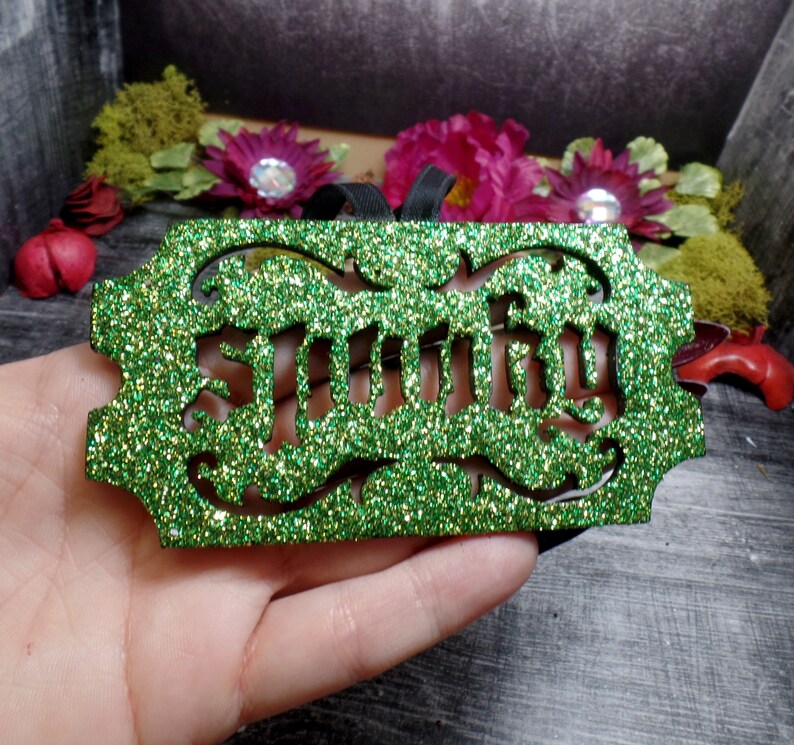 Puede incluir: Un adorno verde brillante con la palabra "Smitty" recortada en una fuente decorativa. El adorno tiene una cinta negra para colgar y est&aacute; rodeado de flores y vegetaci&oacute;n artificiales.