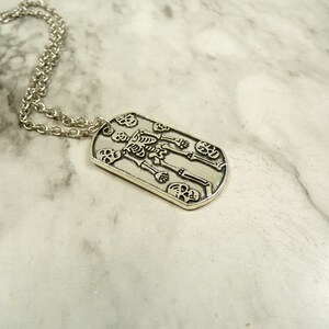 May include: Silver necklace with a dog tag pendant featuring a skeleton design with skulls.
