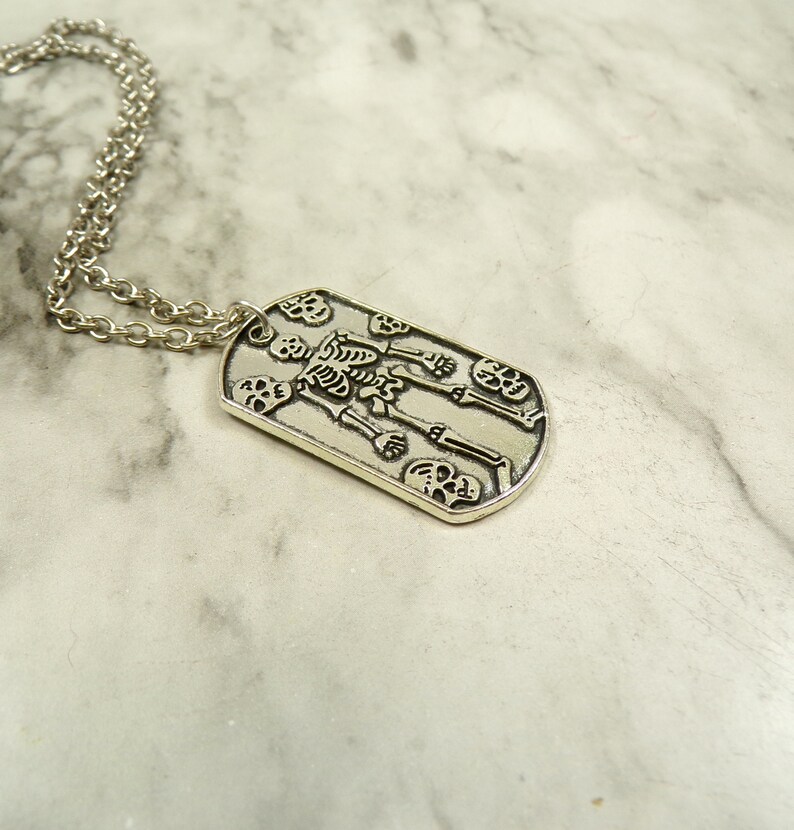 May include: Silver necklace with a dog tag pendant featuring a skeleton design with skulls.