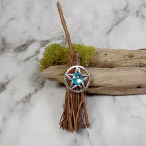 May include: A small, handmade broom with a brown twig handle and a bundle of brown twigs for the bristles. A silver pentagram charm with a blue gemstone in the center is attached to the handle.