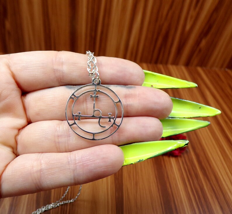 May include: A silver necklace with a circular pendant featuring a sigil with a cross and other symbols. The pendant is on a silver chain.