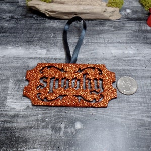 May include: An orange glitter ornament with the word "Spooky" cut out. It has a dark grey ribbon for hanging. A coin is next to the ornament for size comparison. The ornament is on a grey wooden surface.