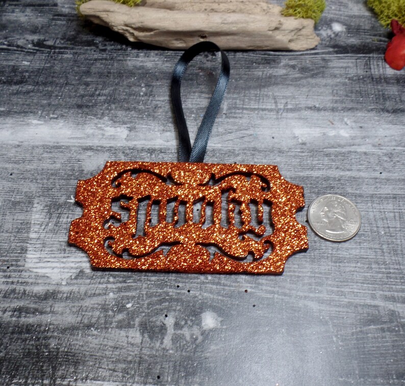 May include: An orange glitter ornament with the word "Spooky" cut out. It has a dark grey ribbon for hanging. A coin is next to the ornament for size comparison. The ornament is on a grey wooden surface.
