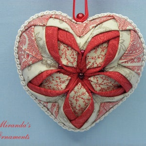 May include: A red and cream heart-shaped fabric ornament with a white trim. The ornament is made of quilted fabric and has a flower design in the centre.