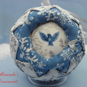 May include: A handcrafted Christmas ornament with a blue and white snowflake pattern. The ornament features a central design with a blue bird and is accented with a silver ribbon for hanging. The text "Miranda's Ornaments" is visible.