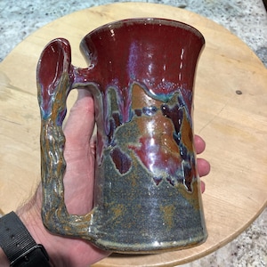 May include: A handcrafted ceramic mug with a unique handle design. The mug features a rich, glossy glaze in shades of burgundy, brown, and blue. The handle has a textured, organic appearance. This mug is perfect for coffee or tea.