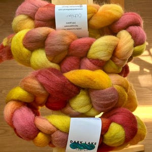 May include: A braided hank of 100% Corriedale wool fiber in a variegated color palette of yellow, orange, and deep red. The label reads October, 100 grams, nonsuperwash, and Dark Lake fiber art with a logo of a teal wave design.