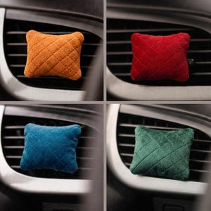 May include: A set of four small, rectangular car air freshener pillows with a quilted texture. The pillows are available in orange, red, blue, and green colours. Each soft, plush pillow is designed to be attached to a vehicle's air vent.