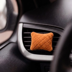 Op de afbeelding: Een kleine, rechthoekige, oranje auto-luchtverfrisser met een gewatteerd ruitpatroon. Het item is bevestigd aan de horizontale lamellen van een zwart ventilatierooster en heeft een zacht, pluche-achtig oppervlak.
