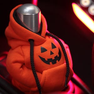 May include: An orange hoodie with a black jack-o'-lantern face design. The hoodie has a black drawstring and is placed over a silver object. The background is dark with red lighting, creating a Halloween theme.