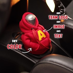 May include: A red hoodie-shaped car gear shift cover with a yellow "A" on the front. The cover has a silver top and red drawstrings. The image also includes text that says "ANY TEAM LOGO", "ANY IMAGE", "ANY TEXT", and "ANY COLOR".