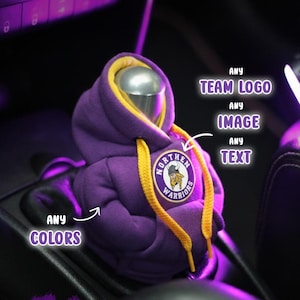 May include: A purple hoodie-shaped car gear shift cover with yellow drawstrings and a patch that reads "Northern Warriors." The image also includes the text "Any Colors," "Any Team Logo," "Any Image," and "Any Text."