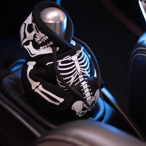 May include: Black hoodie-style shift knob cover featuring a white skeleton design. The cover has a drawstring and is placed over a silver shift knob in a car interior. The design includes a skull and rib cage.