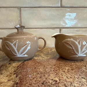 May include: A brown ceramic creamer and sugar bowl set with a white leaf design. The creamer has a handle and the sugar bowl has a lid.