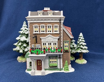 May include: A miniature brick building with white trim and a red roof. The building has a sign that reads "The Crown & Cricket". The building is decorated with greenery and red bows for the festive season.