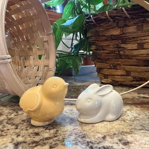 May include: A yellow ceramic chick and a white ceramic bunny figurine sit on a speckled surface. A light brown woven basket and a darker brown woven basket are in the background. Green plants are also visible.
