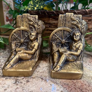 Puede incluir: Un par de sujetalibros dorados antiguos. Cada uno presenta una figura sentada leyendo, con una rueda de carro y espigas de trigo. Los sujetalibros son de metal y tienen un diseño detallado y texturizado. Altura aproximada de 15 cm.